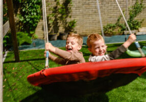 Tweeling Owen en Jake op de nestchommel, vol energie en beweging. De dynamiek in het beeld laat voelen hoe ze zich door de lucht laten dragen, een moment van pure vrijheid en plezier, vastgelegd als onderdeel van een authentiek zorgverhaal thuis.